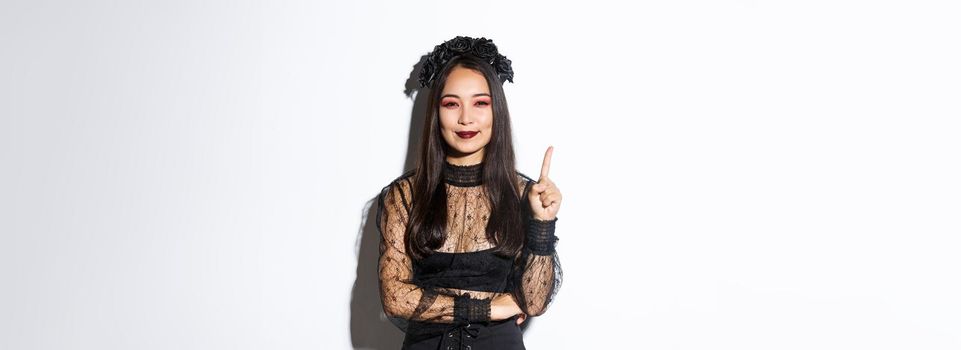 Creative young woman in witch costume smiling pleased as have great idea, raising finger to say suggestion. Female asian dressed-up like widow or mysterious magician, white background.