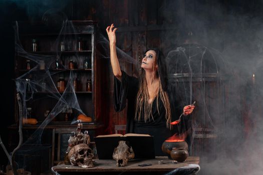Halloween, witch use magic book and cauldron prepare poison or love potion. Black magic occult female wizard in dark room cage spider web human skull. Necromancer Deal with the Devil