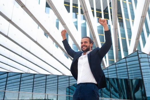 Excited businessman in suit celebrating victory arms raised up. Hispanic male business person winner pose. Best deal ever. Business man expressing positivity standing outdoors office building