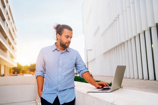 Hispanic businessman standing outside with laptop working remote distant job outdoors near office company building. Stylish unshaved male business man with long hair dressed use laptop