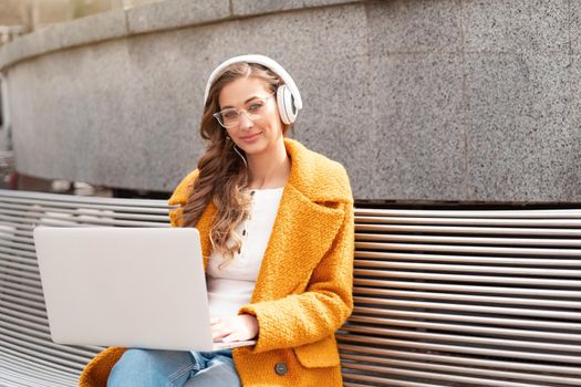 Business Woman Wear Eyeglasses Listen Music Headphone Outdoor Sitting Bench Using Laptop Outdoor Dressed Stylish Yellow Coat Smile Caucasian Female 30s Enjoy Podcast Or Audio Books Outside