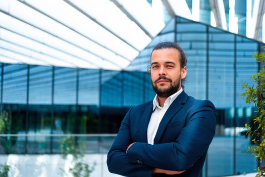 Confidence successful businessman or worker in suit with beard standing in front of office glass building arm crossed. Hispanic handsome business man looking at camera. Portrait shoot. Bank worker