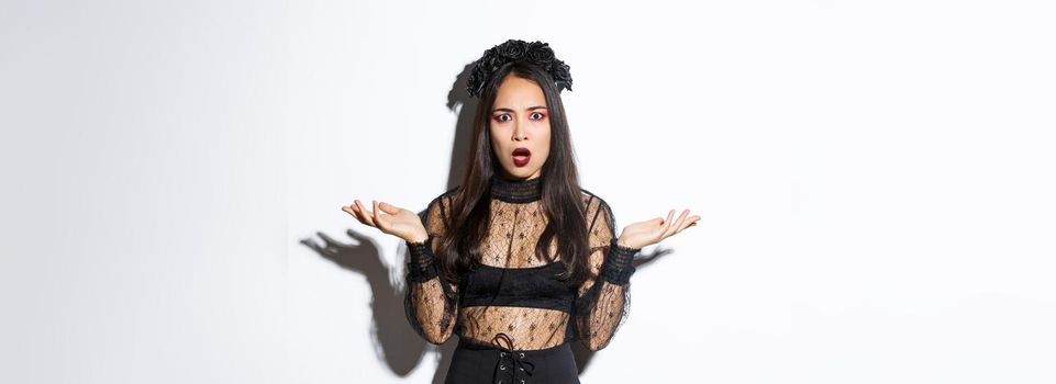 Confused asian woman in halloween costume looking clueless, raising hands sideways and stare puzzled at camera. Female wearing black lace dress, being a witch or magician on trick or treat party.