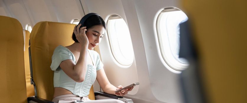 Young woman with mobile phone and headphones listening to music in airplane during flight.