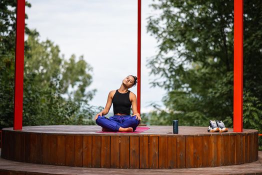 Young woman is practising yoga sitting in the lotus position on a wooden podium in the garden