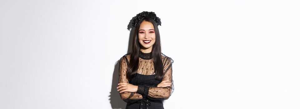 Smiling beautiful asian woman celebrating halloween, wearing black wreath and dress, gothic makeup, looking happy at camera. Female celebrating autumn event, trick or treating in costume.