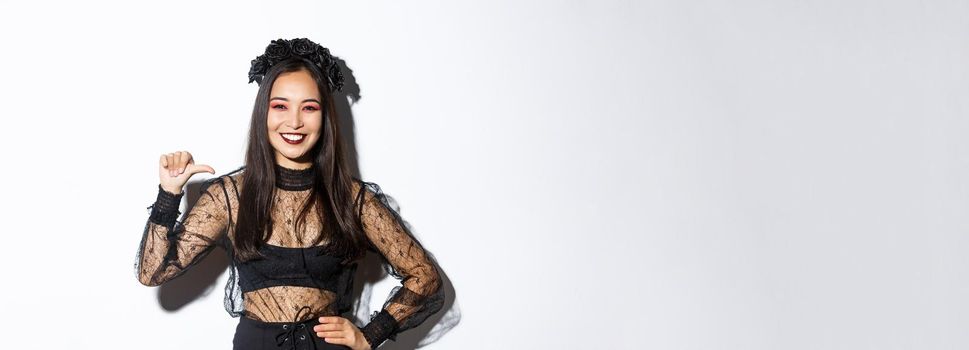 Confident smiling woman feeling like professional, pointing at herself while standing over white background in black gothic dress for halloween party.