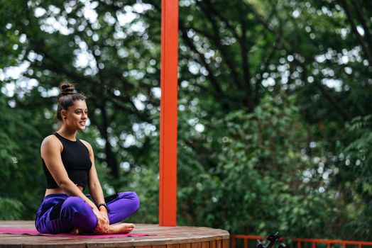 Young woman is practising yoga sitting in the lotus position on a wooden podium in the garden