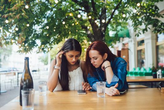 Two female friends browsing in mobile phones while sitting together at city street cafe.