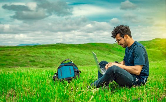 Smiling freelancer young man with laptop working from the field. Close up of man sitting on green grass working with his laptop. Smiling man with laptop sitting on the grass in the field
