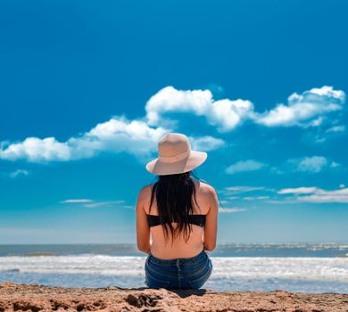 Young woman in hat sitting with her back looking at the sea, vacation girl concept, Girl in hat sitting with her back facing the sea, rear view of a girl looking at the sea