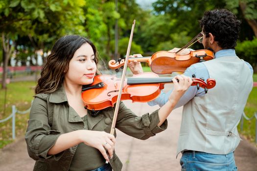 Two young violinists standing playing violin in a park. Portrait of man and woman together playing violin in park. Violinist man and woman back to back playing violin in a park outdoors