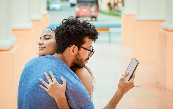 Unfaithful man looking at the cell phone while hugging his girlfriend outdoors. Unfaithful boyfriend hugging his girlfriend and looking at the cell phone. Concept of unfaithful man using cell phone