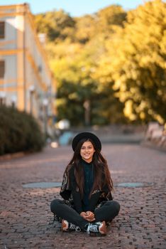 Young woman in hat sitting at city on cobblestone, warm autumn day