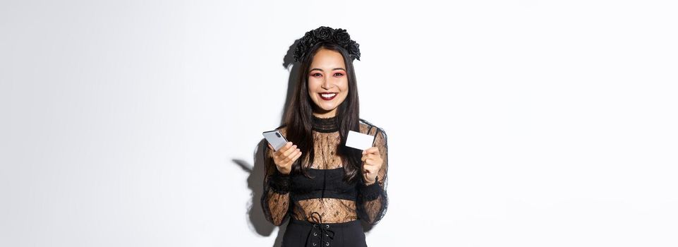 Beautiful stylish asian woman in halloween costume holding mobile phone with credit card and smiling, standing over white background.