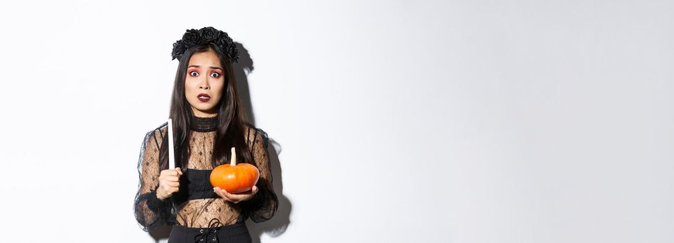 Image of asian woman panicking, being scared of witchcraft on halloween, standing in witch costume with candle and pumpkin.