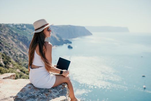 Successful business woman in yellow hat working on laptop by the sea. Pretty lady typing on computer at summer day outdoors. Freelance, travel and holidays concept.