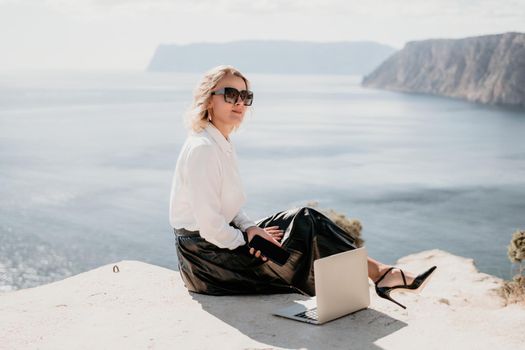 Successful business woman in yellow hat working on laptop by the sea. Pretty lady typing on computer at summer day outdoors. Freelance, travel and holidays concept.