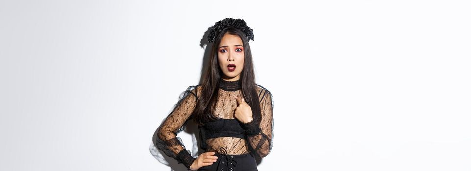 Insulted and confused asian woman in halloween costume pointing at herself, wearing black dress of evil witch on party, standing over white background.