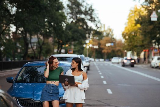 Two young women with a laptop near the car on the road