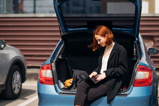 Business attractive young woman working on laptop while sitting in trunk of car while traveling