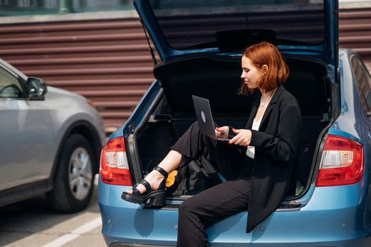 Business attractive young woman working on laptop while sitting in trunk of car while traveling