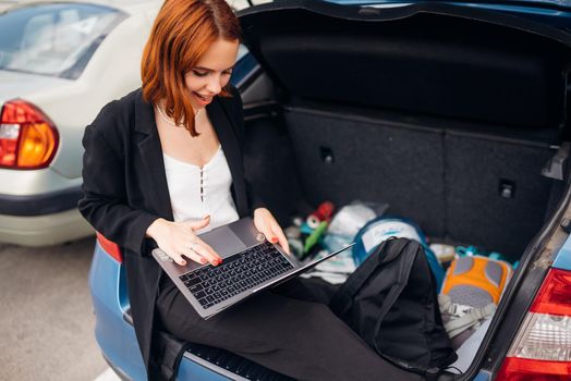 Business attractive young woman working on laptop while sitting in trunk of car while traveling