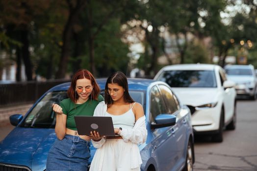 Two young women with a laptop near the car on the road