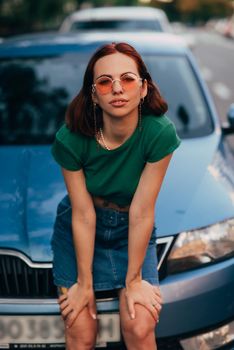 Beautiful girl in sunglasses sits on the hood of a blue car, against the road