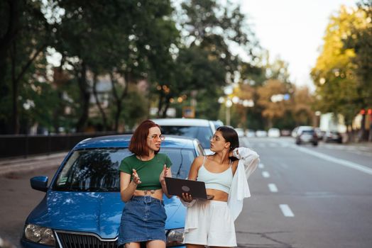 Two young women with a laptop near the car on the road