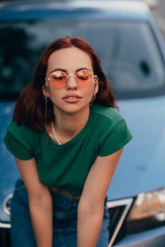 Beautiful girl in sunglasses sits on the hood of a blue car, against the road