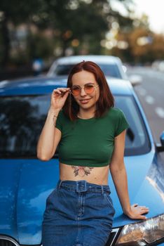 Beautiful girl in sunglasses sits on the hood of a blue car, against the road