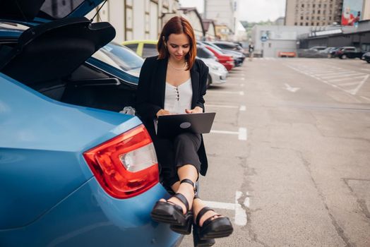 Business attractive young woman working on laptop while sitting in trunk of car while traveling