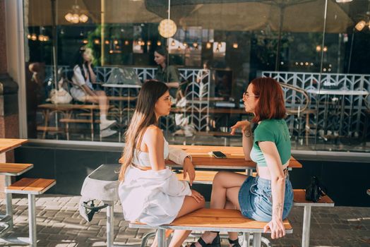 Two pretty girlfriends talking while sitting in a bar at the city street
