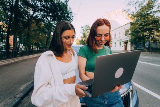 Two young women with a laptop near the car on the road