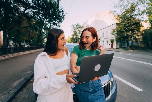 Two young women with a laptop near the car on the road
