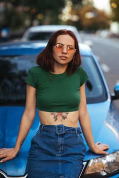 Beautiful girl in sunglasses sits on the hood of a blue car, against the road