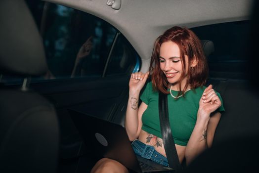 Beautiful woman is using a laptop and smiling while sitting on back seat in the car