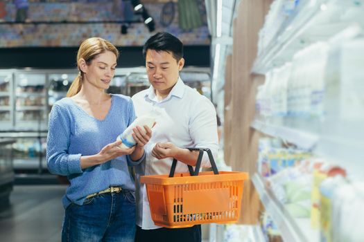 Family shopping. Young interracial couple, Asian man and woman choosing milk in supermarket. They hold a basket and a bottle of milk in their hands, read the label, standing near the refrigerator.