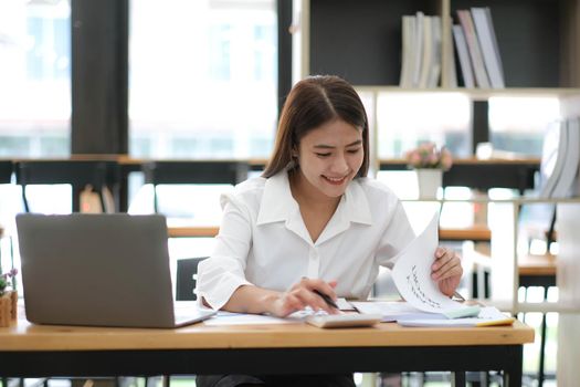 Smiling beautiful Asian businesswoman analyzing chart and graph showing changes on the market and use calculator at office..