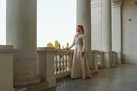 a beautiful mature woman in a long white dress stands on the balcony and looks into the distance.