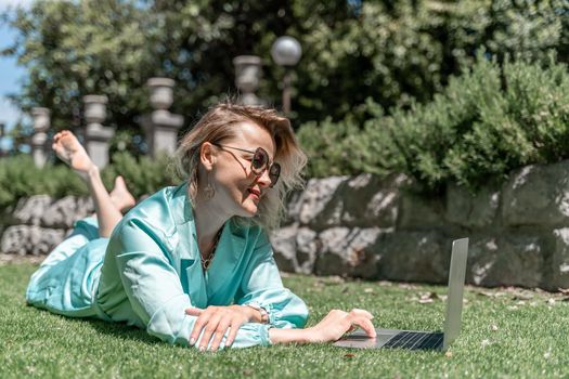 a young beautiful woman with blond curly hair in glasses and a blue dress sits on the grass in nature and uses a laptop