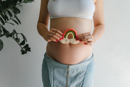 A pregnant girl in a denim jumpsuit holds a rainbow in her hands for the decor of a children's room. Great time to have a baby. Future mom. Rainbow macrame.