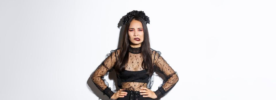 Annoyed and bothered evil witch looking complicated and biting lower lip, standing over white background in black lace dress and wreath. Girl in halloween costume trick or treating.