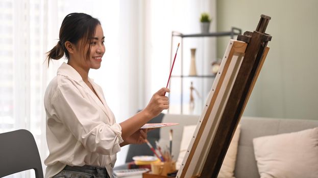 Satisfied young woman painting with watercolor on canvas in bright living room. Art, creative hobby and leisure activity concept.