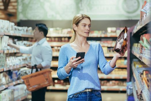 A beautiful young blonde woman is standing in a supermarket. She is holding a phone and a packet of breakfast cereal. Chooses, buys, reads the label. Shoppers in the background.