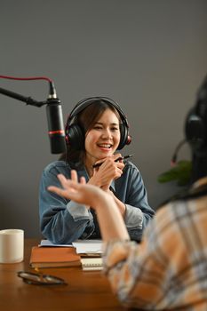 Friendly female radio host looking and talking with her guest while making audio podcast in studio.