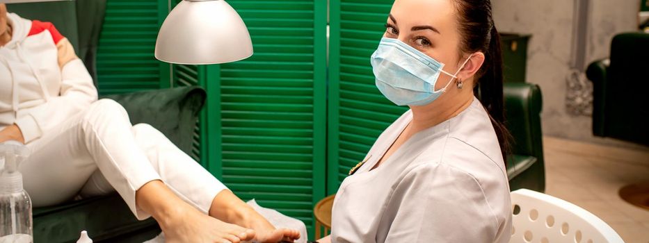 Young female podiatrist with a protective mask doing chiropody and looking at the camera in her podiatry clinic