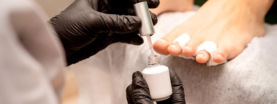 White nail polish in the hands of a manicurist while painting nails on a female feet, closeup