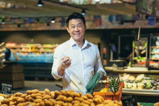 A young handsome Asian man, businessman is standing in a supermarket in the vegetable section, holding a basket of fruits and potatoes in his hands. He looks at the camera, smiles.